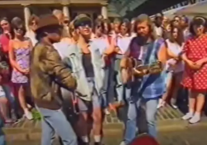 the Bee Gees as they stand in front of an open guitar case and play an acoustic version of their hit songs 'To Love Somebody' and 'Massachusetts' as the crowd stood on all sides watching them sing.